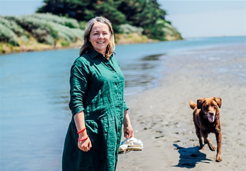 Woman in green dress standing on the beach with her large brown dog running.